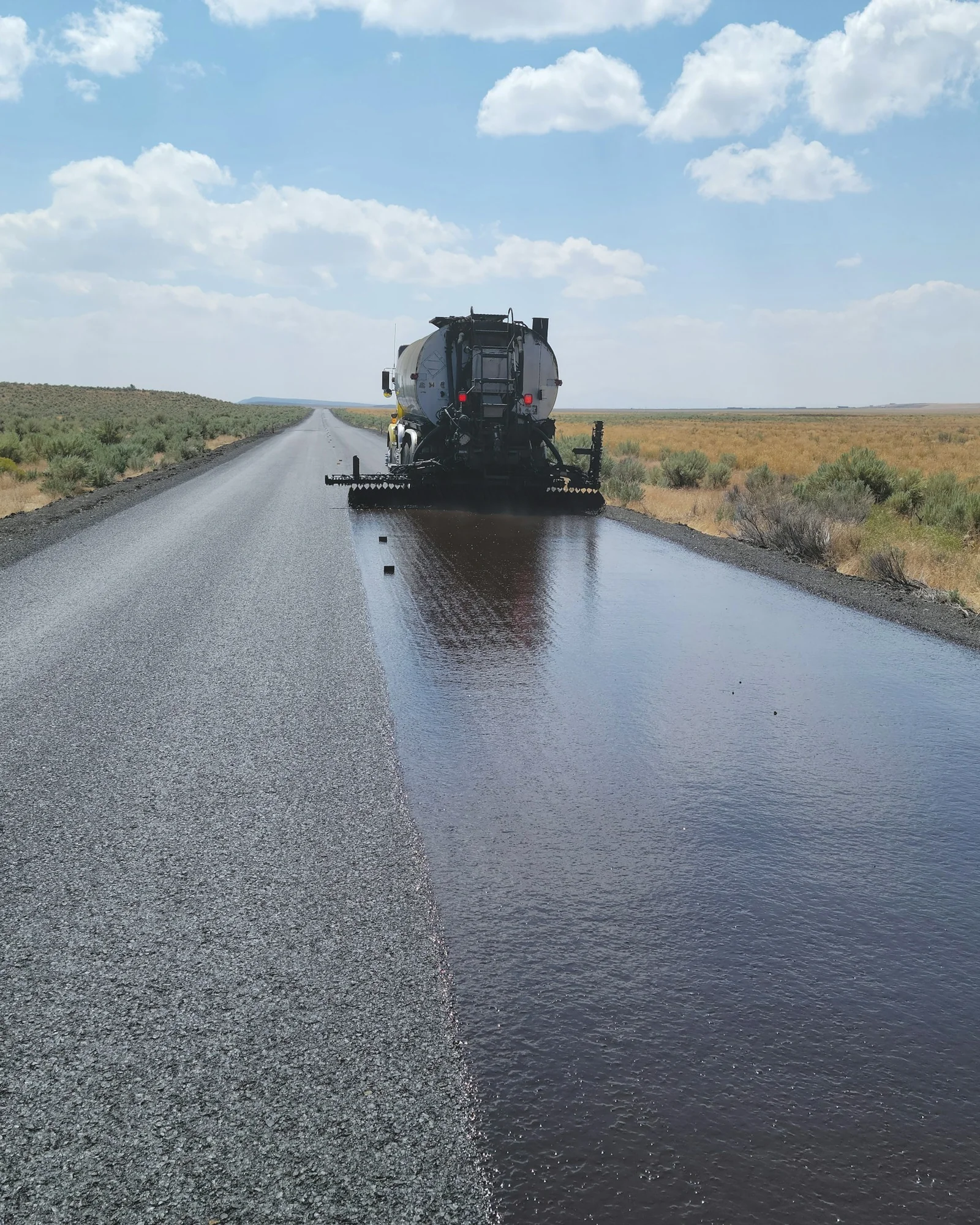 IMG S07 lavori pubblici e Autocisterna che stende bitume su una strada dritta in aperta campagna.