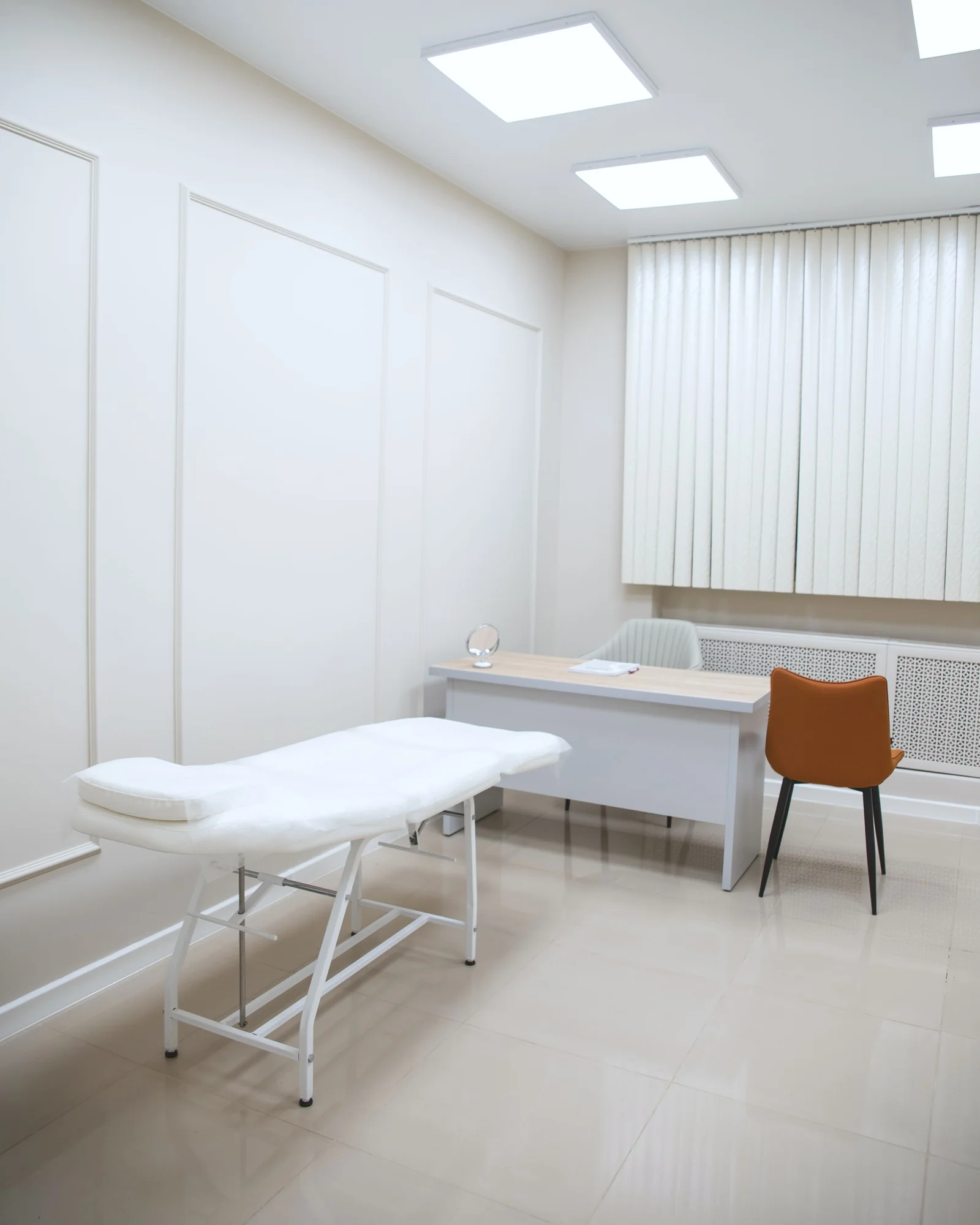 IMG Studio medico con lettino, scrivania, sedia arancione e tende verticali.